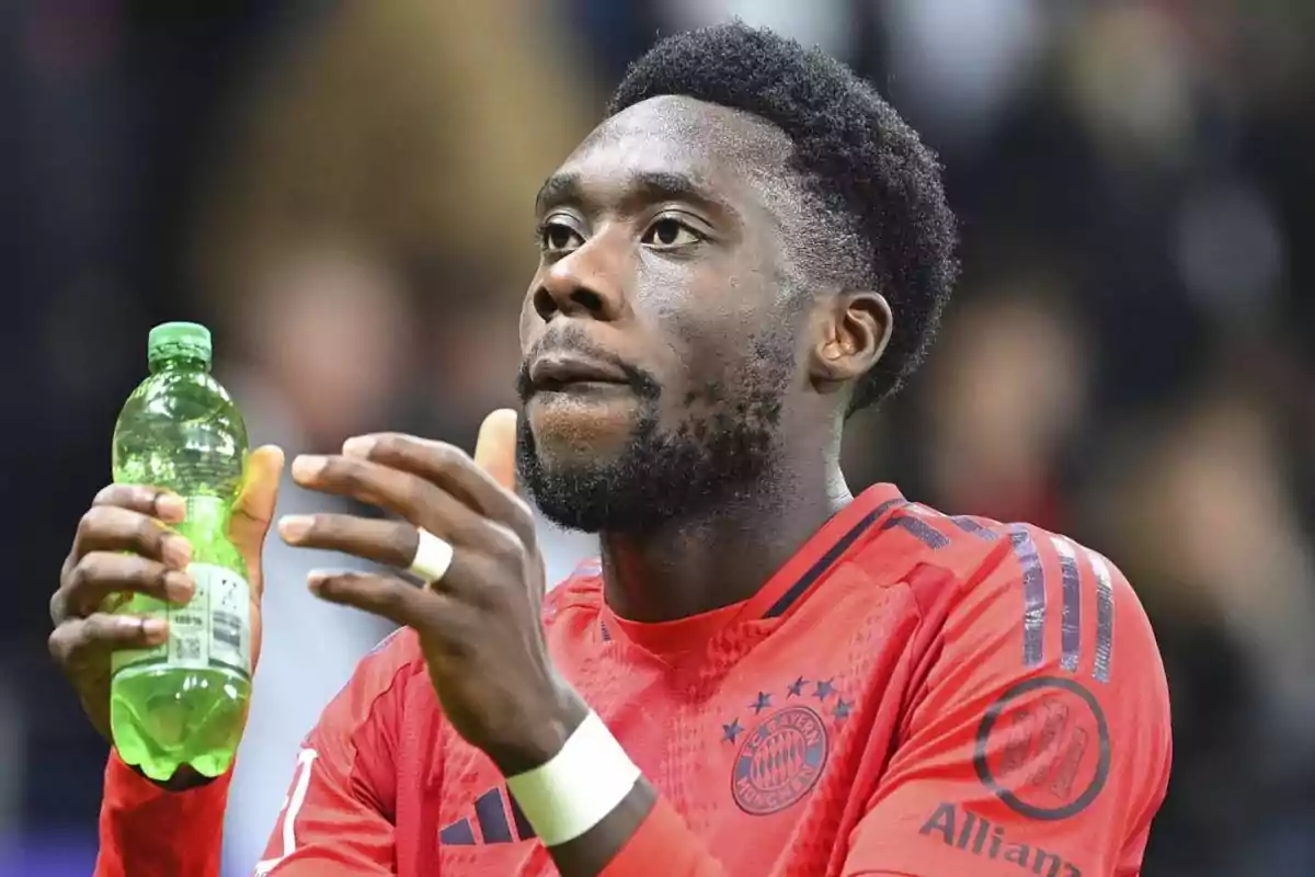 Jugador de fútbol con camiseta roja sosteniendo una botella verde en un estadio.