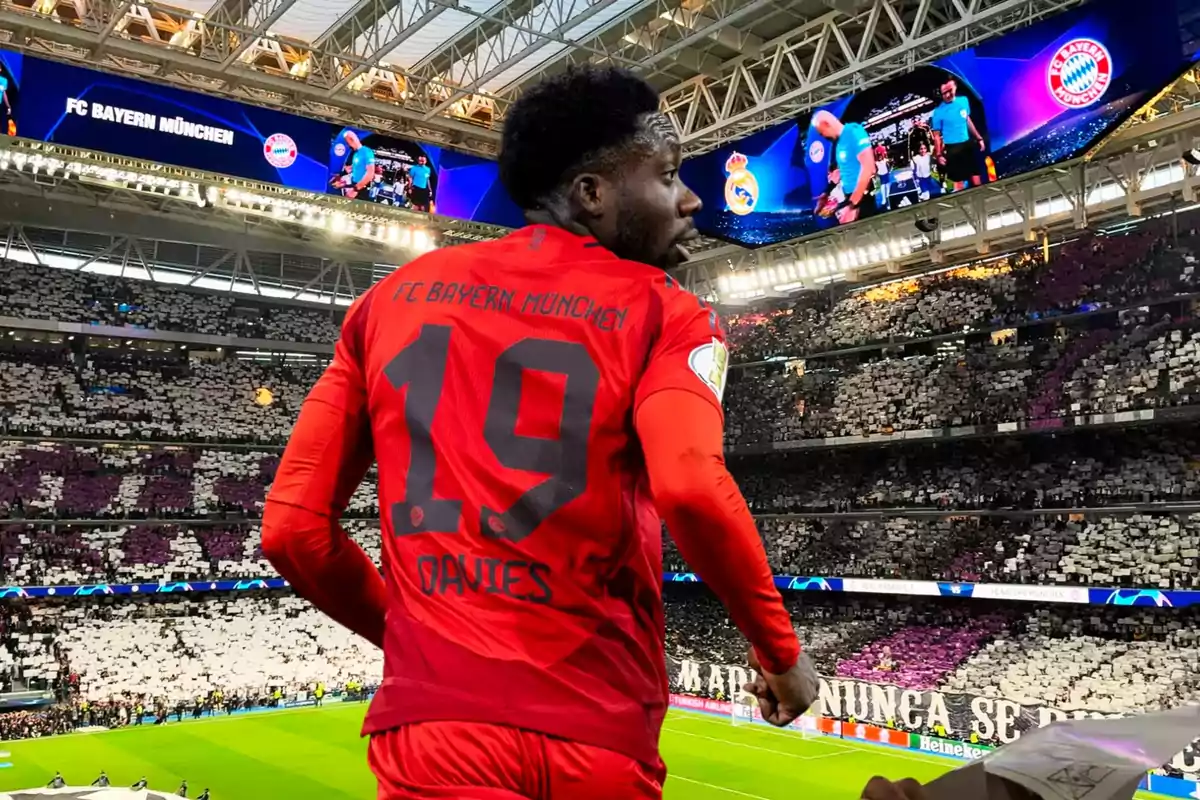 Un jugador del FC Bayern München con el número 19 en su camiseta está en un estadio lleno de espectadores, con una pantalla que muestra el logo del equipo y el de su oponente.