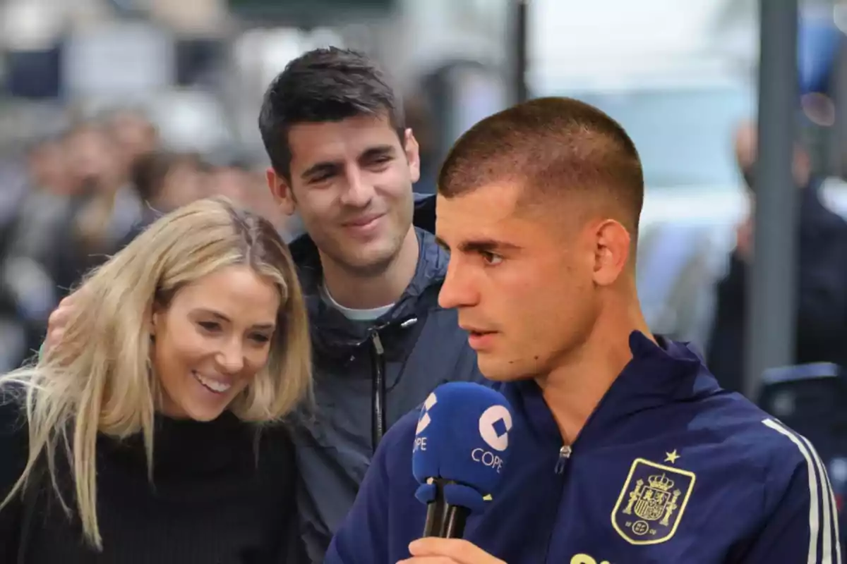 Un hombre con chaqueta deportiva hablando por un micrófono mientras una pareja sonríe en el fondo.