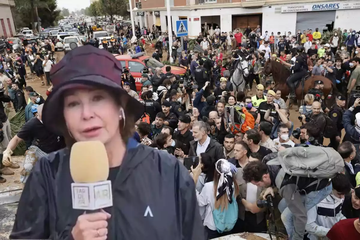 Una reportera con sombrero oscuro sostiene un micrófono mientras una multitud de personas, incluyendo policías y civiles, se congrega en una calle llena de actividad y vehículos.