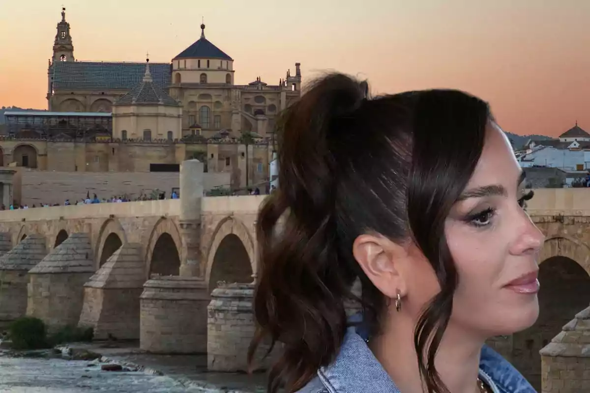 Una mujer de perfil con el cabello recogido frente a un puente histórico y un edificio antiguo al atardecer.