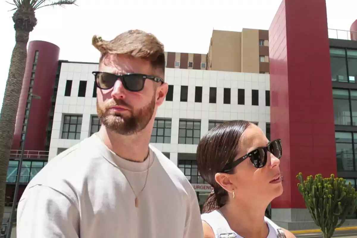 Dos personas con gafas de sol están de pie frente a un edificio moderno con detalles en rojo y blanco, junto a una palmera y un cactus.