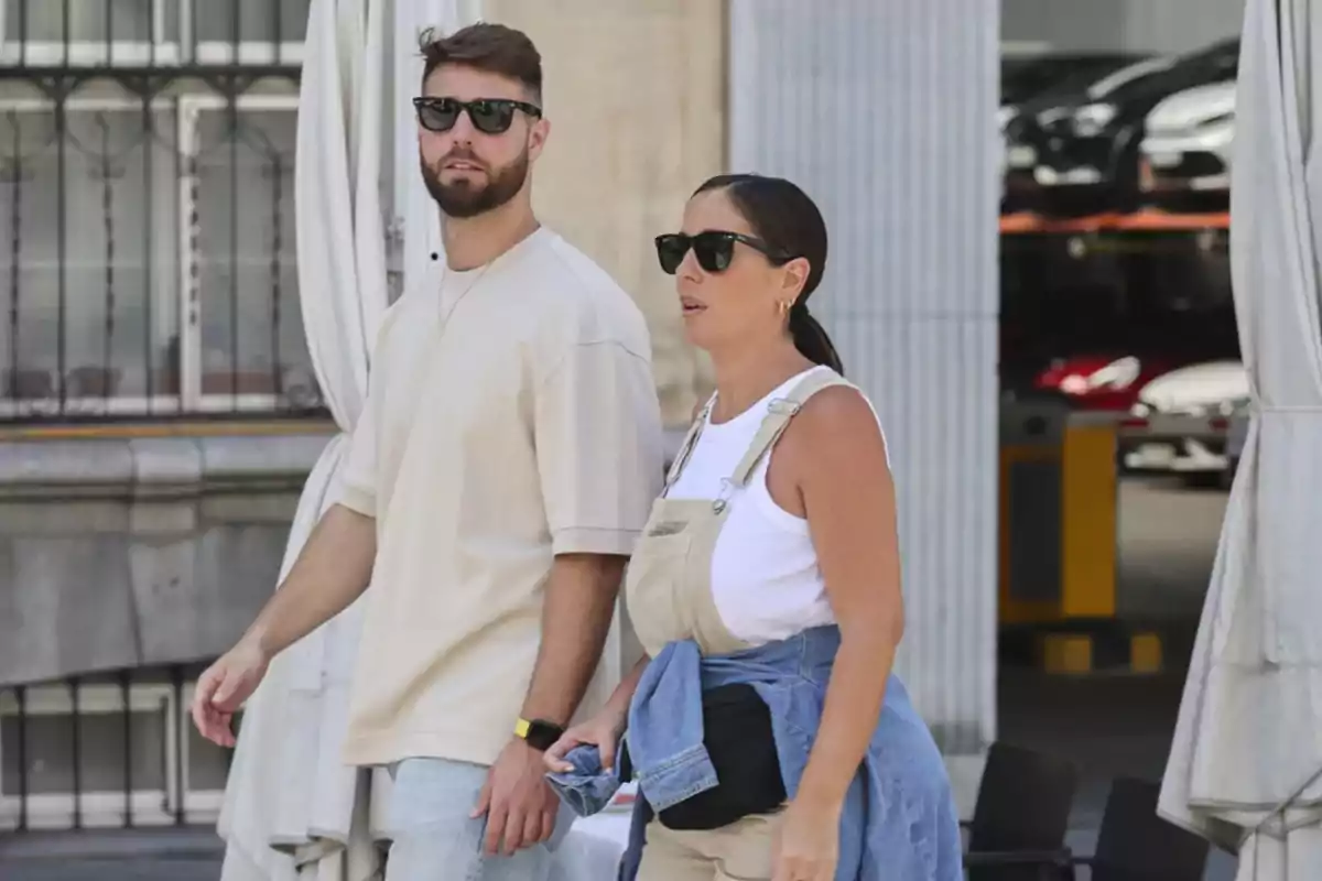 Una pareja caminando por la calle, ambos llevan gafas de sol y ropa casual, el hombre viste una camiseta beige y jeans, mientras que la mujer lleva un mono beige sobre una camiseta blanca y una chaqueta de mezclilla atada a la cintura.