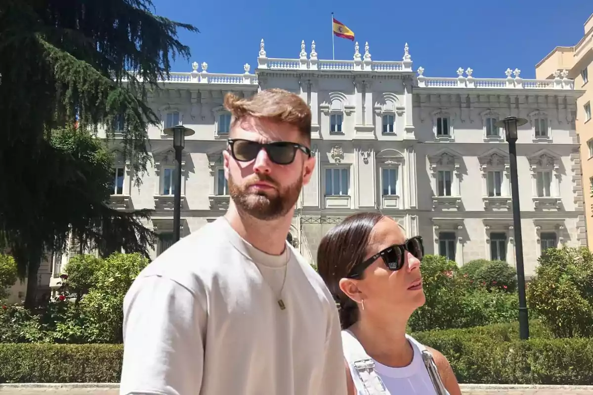 Dos personas con gafas de sol posan frente a un edificio histórico con una bandera española ondeando en la parte superior.