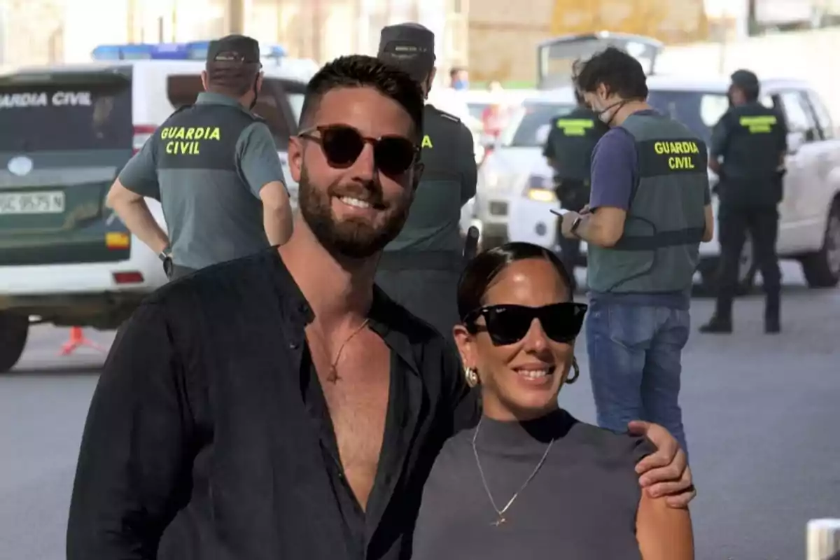 Una pareja sonriente con gafas de sol posa frente a un grupo de agentes de la Guardia Civil y vehículos oficiales.