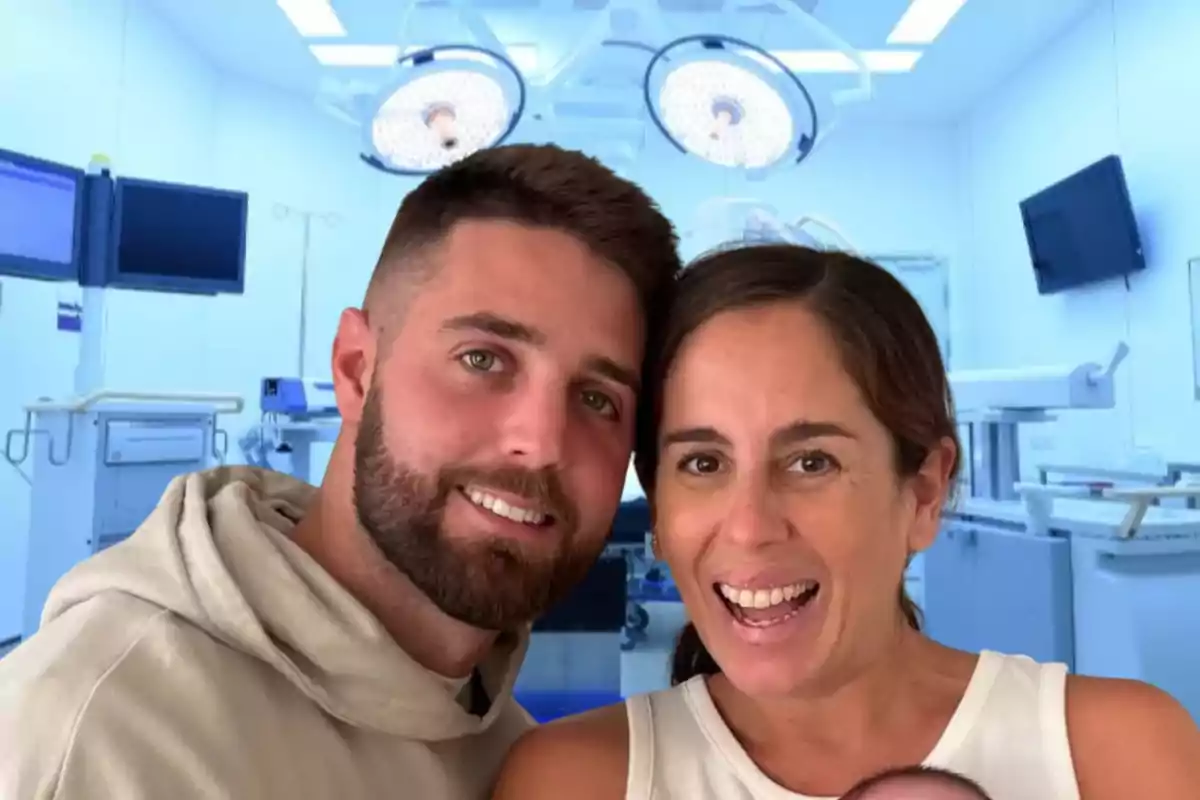 Una pareja sonriente posa en un entorno de hospital con luces quirúrgicas en el techo.