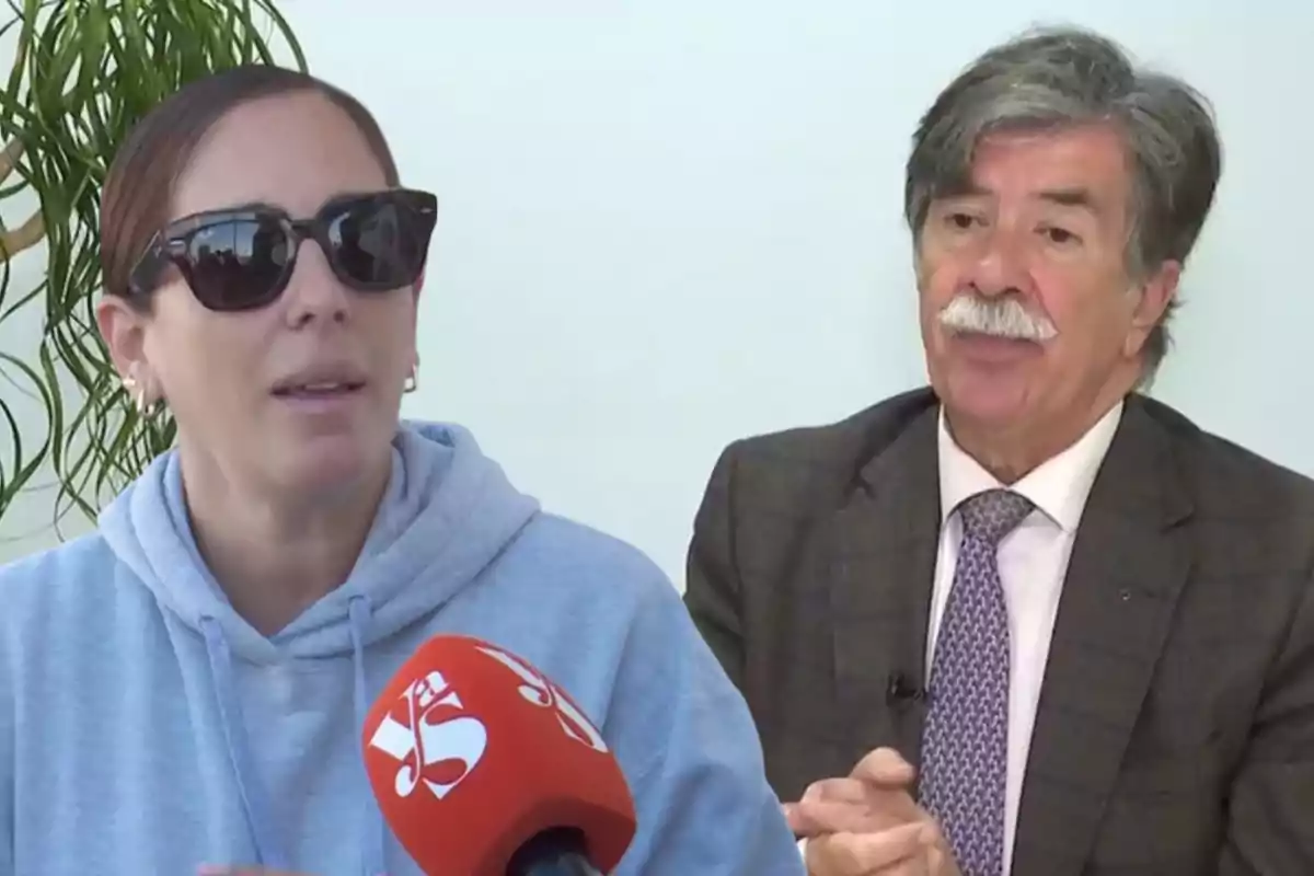 Una mujer con gafas de sol y sudadera azul está hablando frente a un micrófono rojo, mientras un hombre mayor con bigote y traje gris la observa.