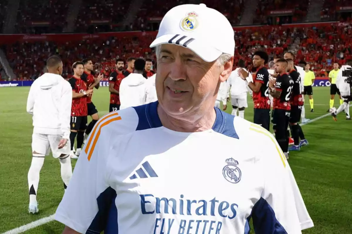 Un entrenador de fútbol con gorra y camiseta del Real Madrid sonríe en un estadio mientras los jugadores de ambos equipos se saludan en el fondo.