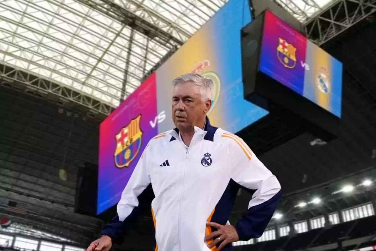 Un entrenador de fútbol con una chaqueta blanca y azul está de pie en un estadio con una pantalla gigante de fondo que muestra los escudos de los equipos FC Barcelona y Real Madrid.