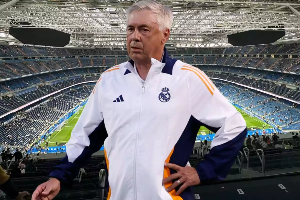 Un hombre con una chaqueta deportiva blanca y azul con el logo del Real Madrid, de pie en un estadio de fútbol vacío.