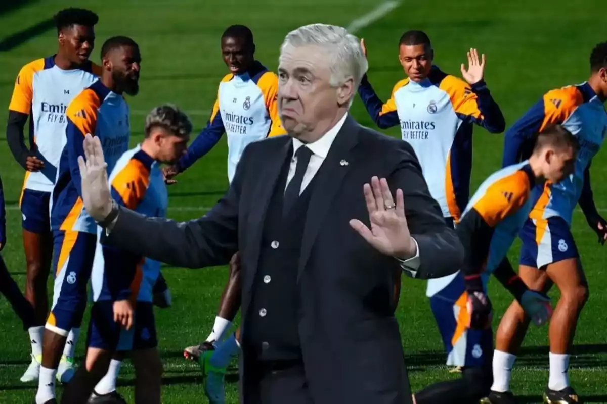 Un entrenador de fútbol con traje hace un gesto de calma con las manos mientras los jugadores entrenan en el campo.