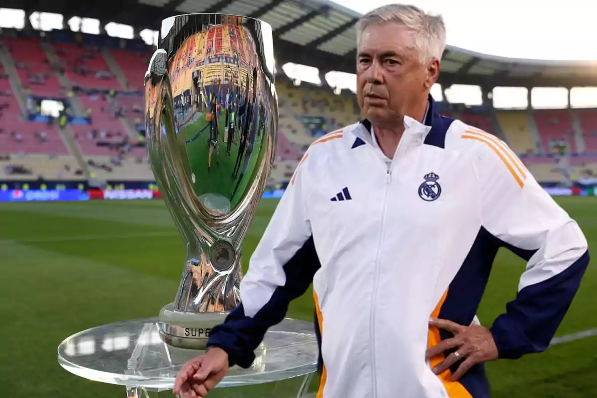 Un entrenador de fútbol con una chaqueta del Real Madrid está de pie junto a un trofeo en un estadio.