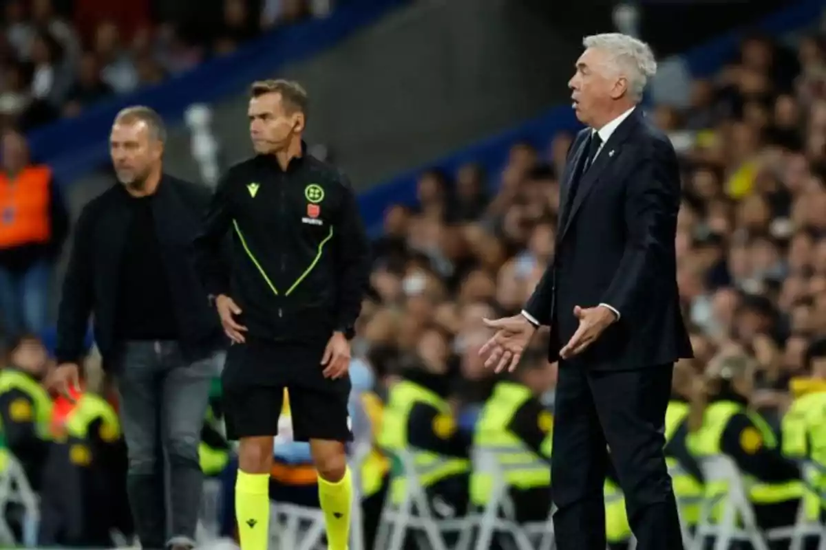 Un entrenador de fútbol gesticula en la línea lateral mientras un árbitro y otro hombre están cerca durante un partido.