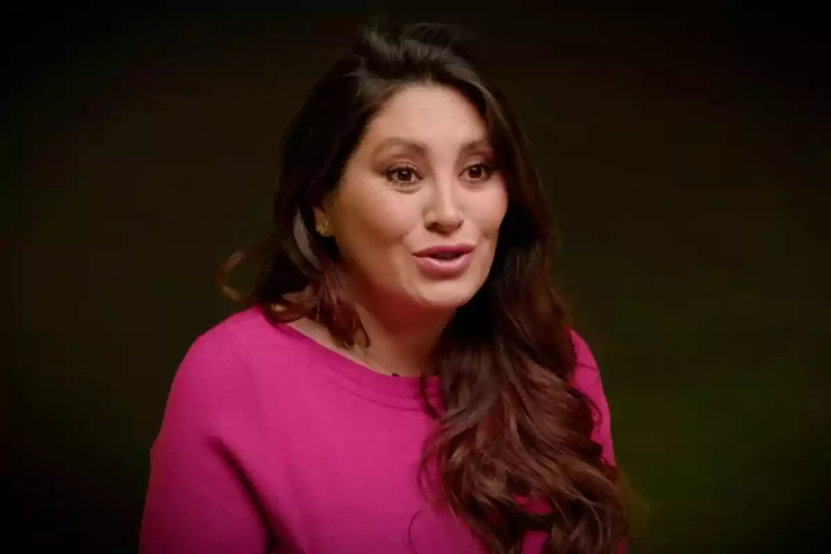 Una mujer con cabello largo y oscuro lleva un suéter rosa y está hablando frente a un fondo oscuro. Una mujer con cabello largo y oscuro lleva un suéter rosa y está hablando frente a un fondo oscuro.