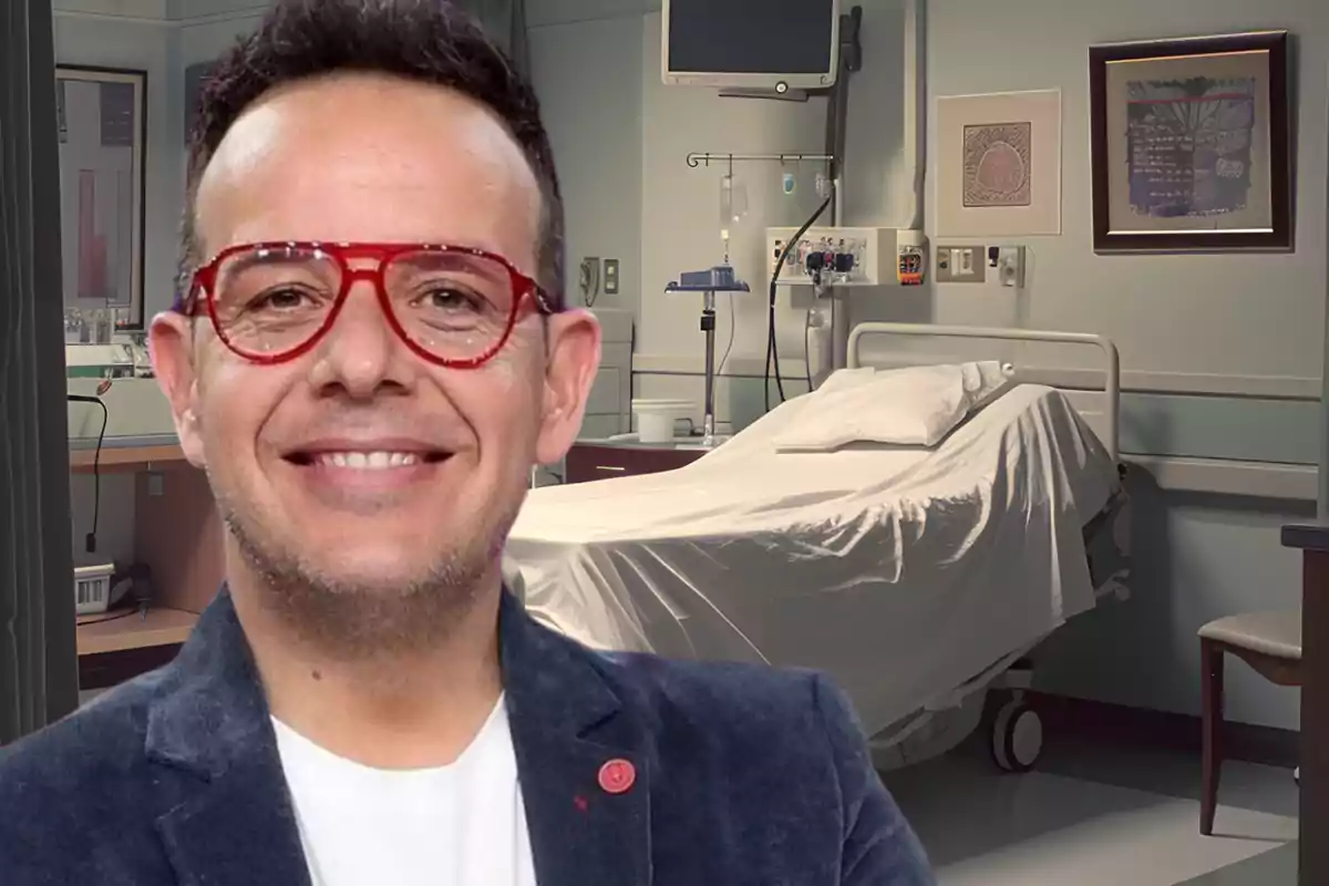 Un hombre con gafas rojas y chaqueta oscura sonríe frente a una cama de hospital.