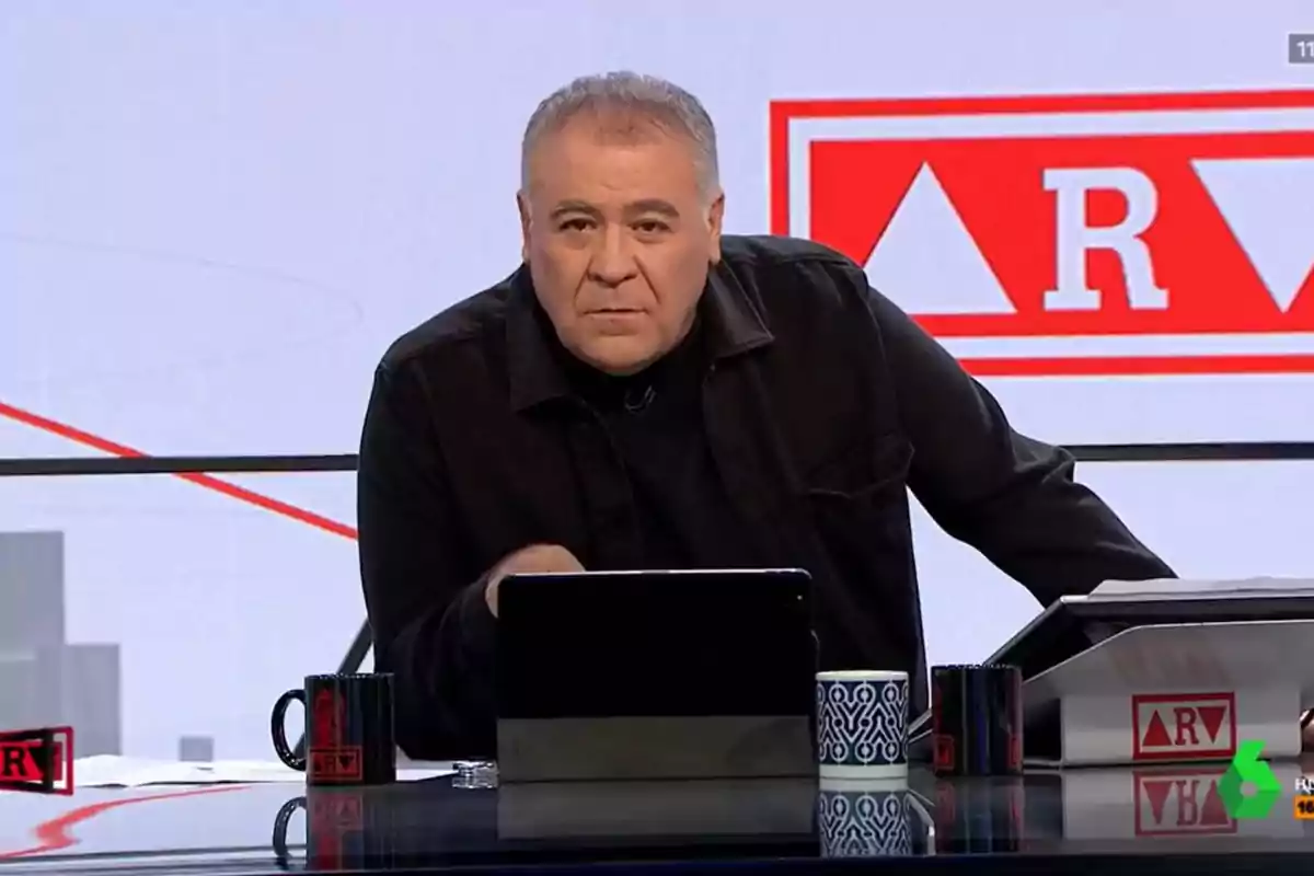 Un presentador de televisión en un estudio con una tableta y tazas sobre la mesa.