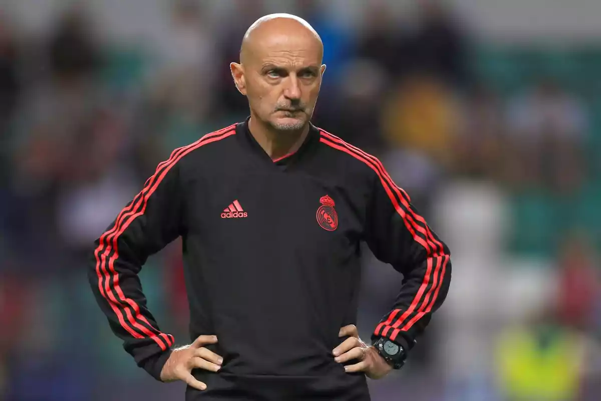 Un hombre calvo con barba incipiente, vestido con un chándal negro con detalles rojos y el escudo de un equipo de fútbol, con las manos en la cintura y una expresión seria.