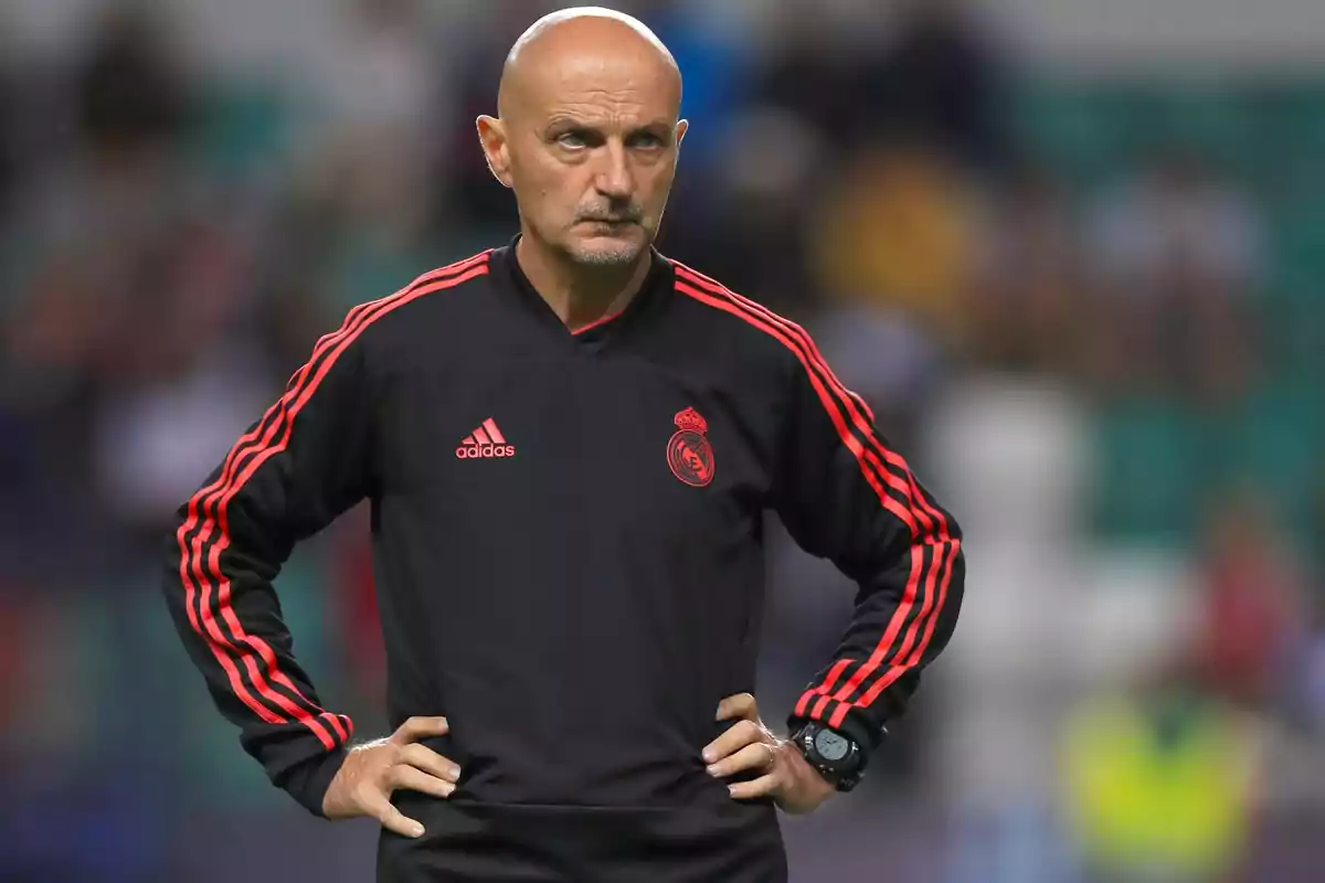 Un hombre calvo con barba incipiente, vestido con un chándal negro con detalles rojos y el logo de Adidas y un escudo de equipo, está de pie con las manos en la cintura en un entorno deportivo.