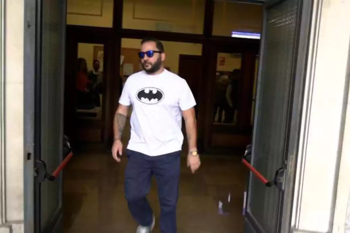 Un hombre con gafas de sol y camiseta con el logo de Batman sale de un edificio a través de una puerta de cristal.