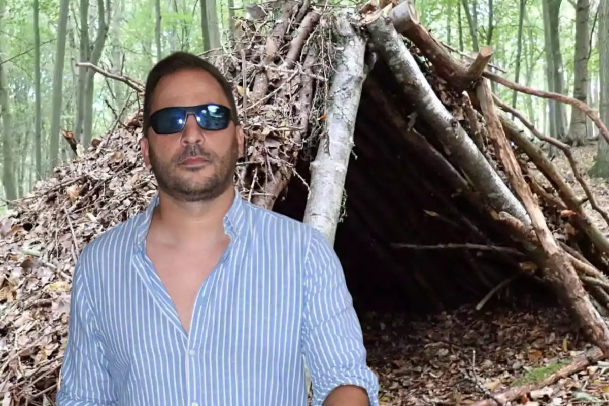 Un hombre con gafas de sol y camisa de rayas azules está de pie frente a un refugio de ramas en un bosque.