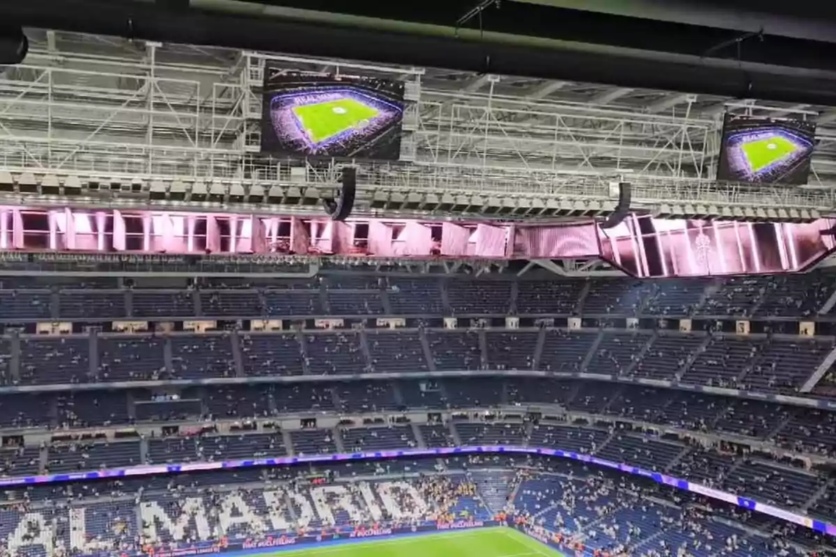 Vista interior de un estadio de fútbol con gradas parcialmente ocupadas y pantallas mostrando el campo iluminado.