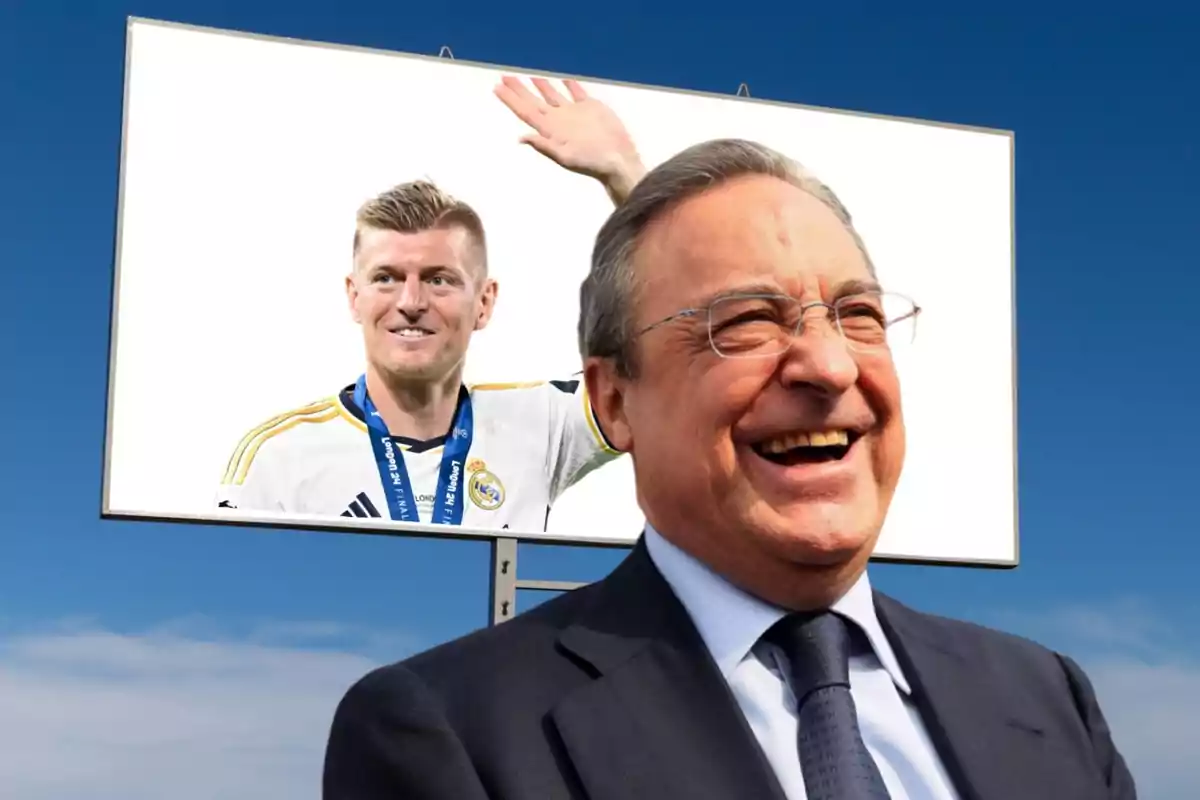 Un hombre sonriente con gafas y traje oscuro está frente a una valla publicitaria que muestra a un jugador de fútbol con una medalla y la camiseta del Real Madrid.