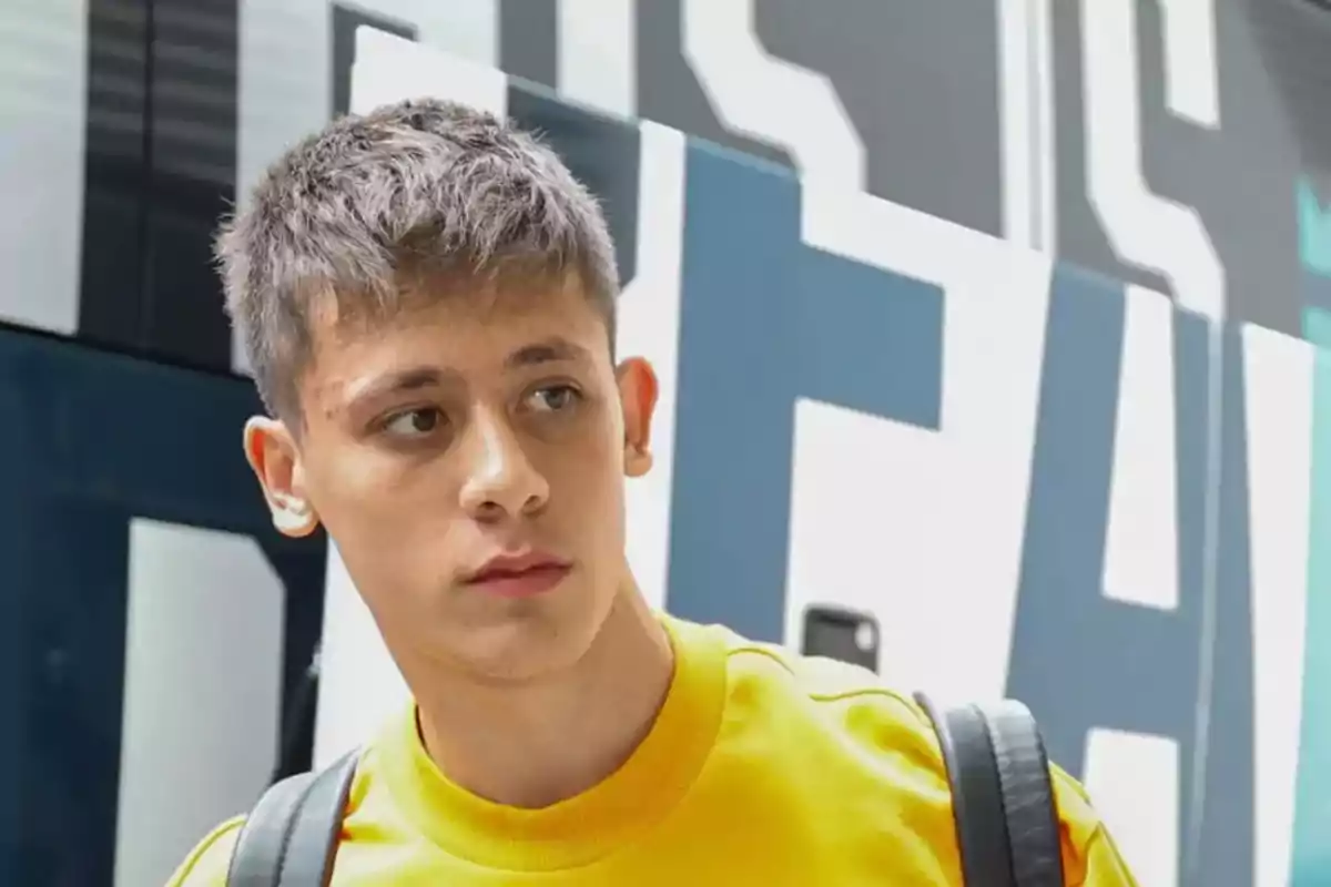 Un joven con camiseta amarilla y mochila negra mira hacia un lado con una expresión seria, con un fondo de letras grandes y borrosas.