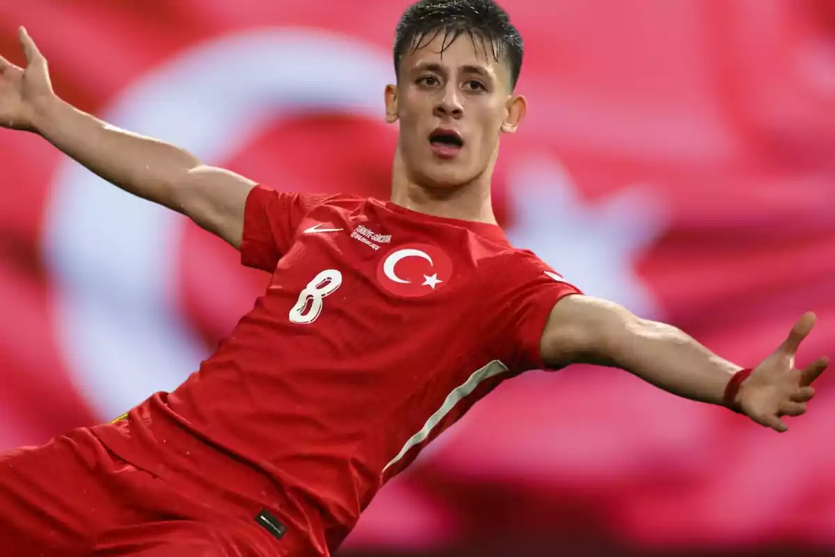 Jugador de fútbol con uniforme rojo celebrando frente a una bandera de Turquía.