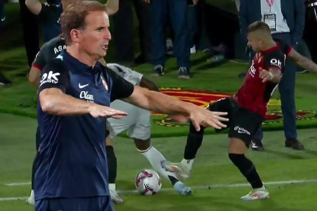 Un entrenador de fútbol gesticulando en la línea de banda mientras dos jugadores disputan el balón en el campo.