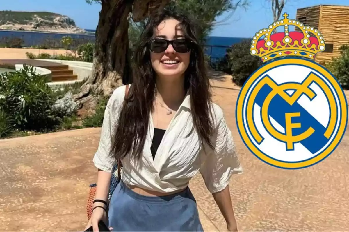 Una mujer sonriente con gafas de sol y una blusa blanca, con el logo del Real Madrid a su derecha, en un entorno al aire libre cerca del mar.