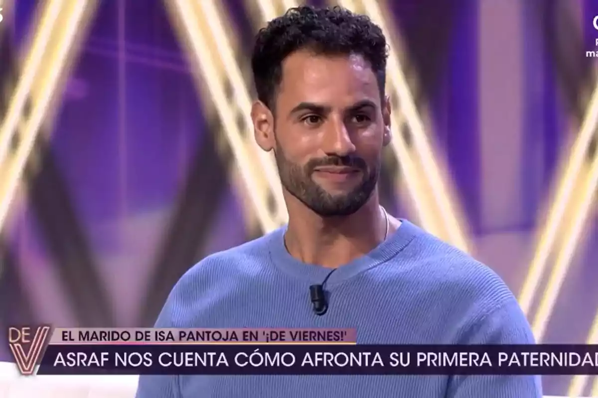 Un hombre con barba y cabello rizado, vestido con un suéter azul, aparece en un programa de televisión con un fondo de luces brillantes y un rótulo que menciona su primera paternidad.