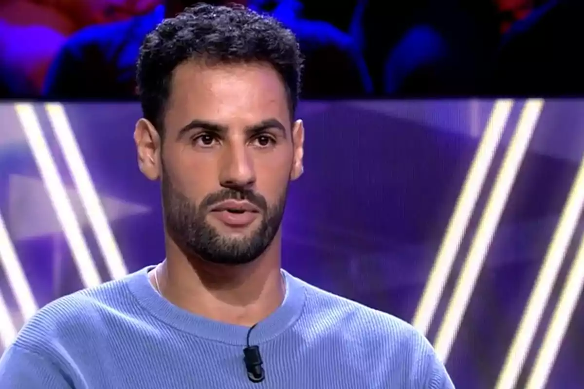 Un hombre con barba y cabello oscuro lleva un suéter azul y un micrófono en un estudio de televisión iluminado con luces de fondo.