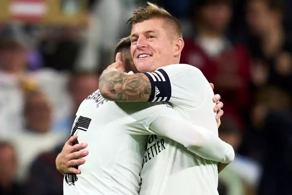 Dos jugadores de fútbol abrazándose en el campo, uno de ellos con un tatuaje en el brazo y una banda de capitán.