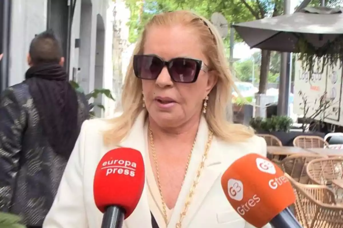 Mujer con gafas de sol y chaqueta blanca siendo entrevistada por medios de comunicación con micrófonos de Europa Press y G3.