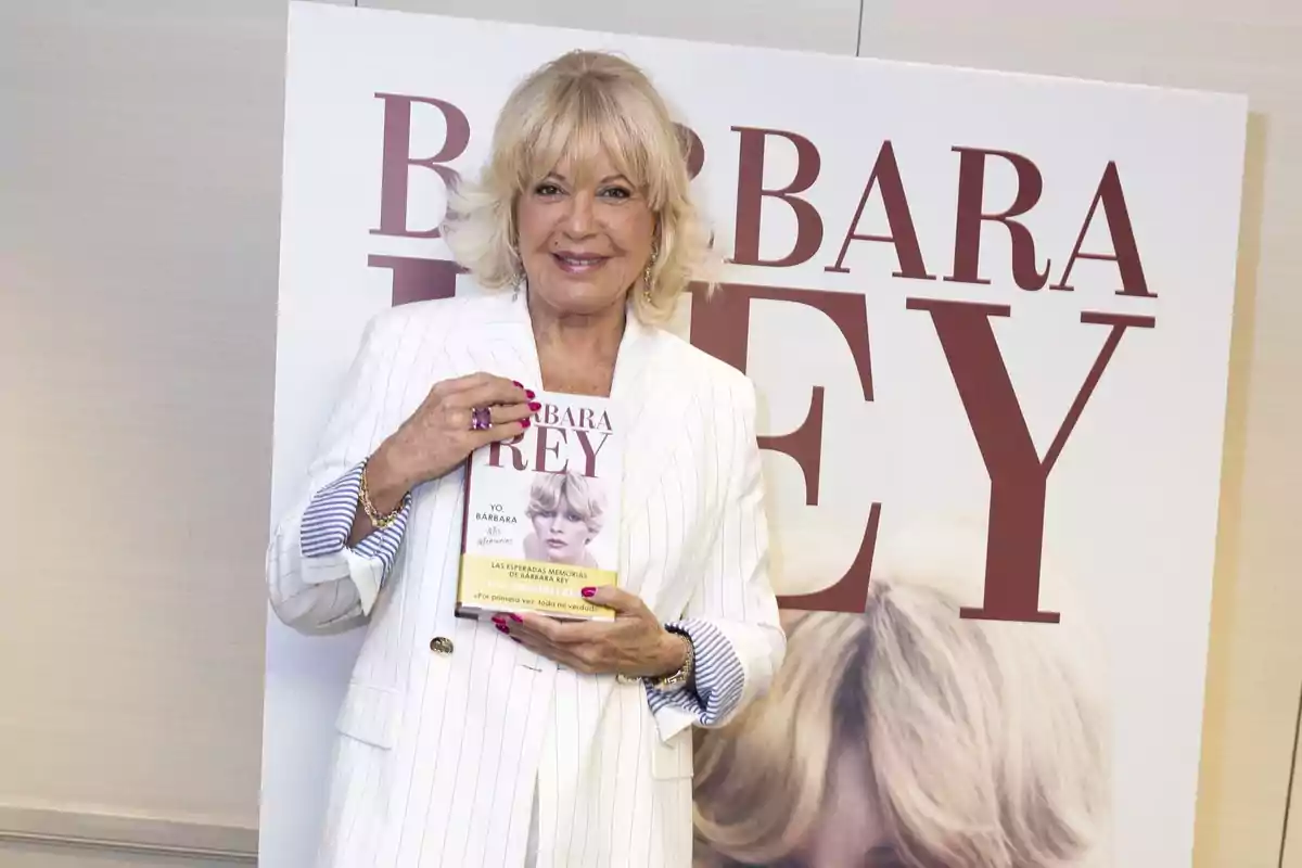 Mujer rubia con saco blanco sostiene un libro frente a un cartel grande con su nombre en letras rojas Mujer rubia con saco blanco sostiene un libro frente a un cartel grande con su nombre en letras rojas