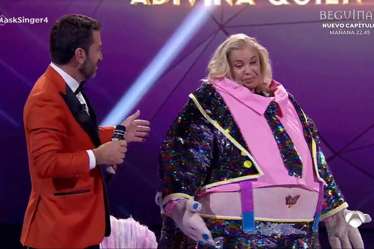 Arturo Valls con chaqueta naranja interactúa con Bárbara Rey disfrazada en un escenario iluminado en Mask Singer. Arturo Valls con chaqueta naranja interactúa con Bárbara Rey disfrazada en un escenario iluminado en Mask Singer.