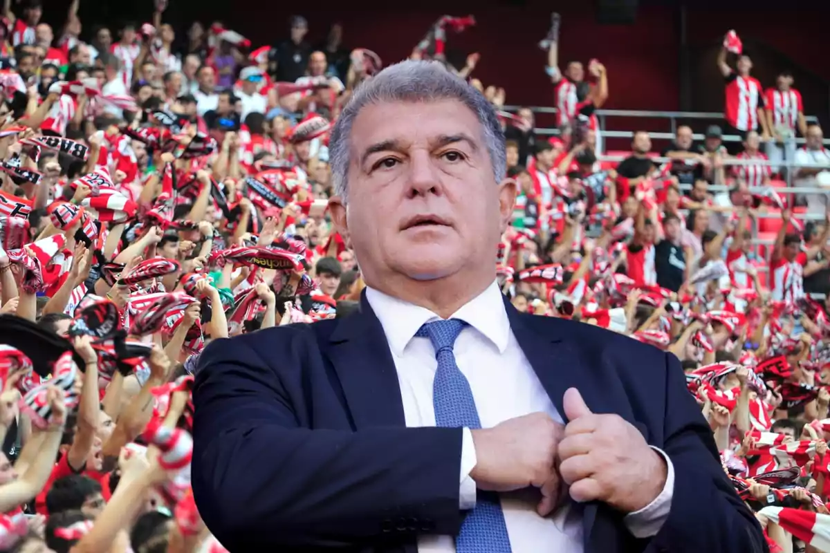 Hombre de traje y corbata frente a una multitud de aficionados con bufandas rojas y blancas en un estadio