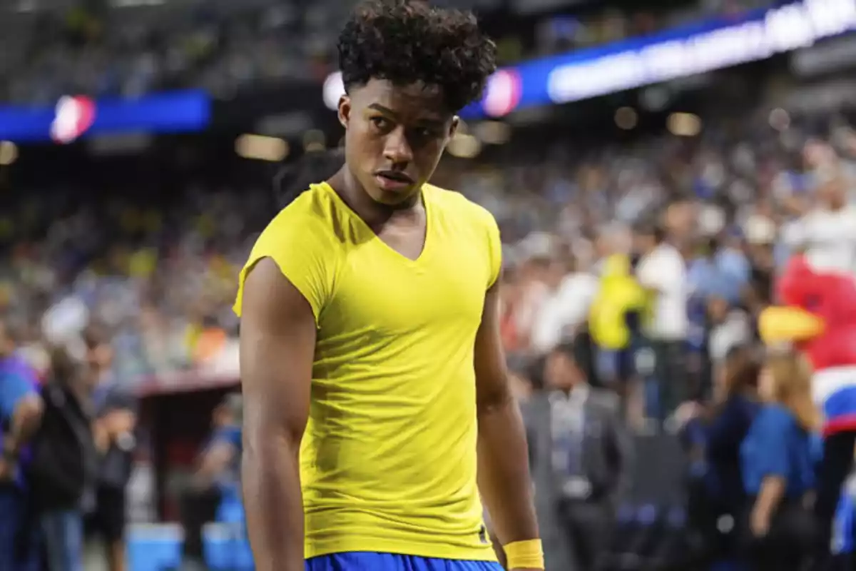 Un joven con camiseta amarilla y pantalones azules está en un estadio lleno de gente.