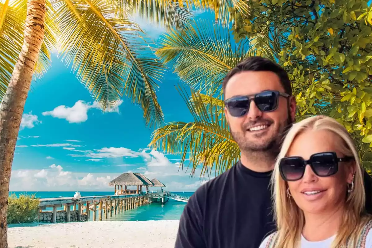 Una pareja sonriente con gafas de sol posa frente a un paisaje tropical con palmeras, una playa de arena blanca y un muelle que se extiende hacia el mar con cabañas sobre el agua.