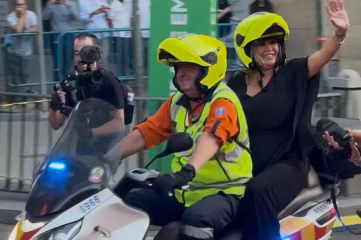 Dos personas con casco amarillo van en una motocicleta mientras una de ellas sonríe y saluda, al fondo hay un camarógrafo y varias personas observando detrás de una valla Dos personas con casco amarillo van en una motocicleta mientras una de ellas sonríe y saluda, al fondo hay un camarógrafo y varias personas observando detrás de una valla