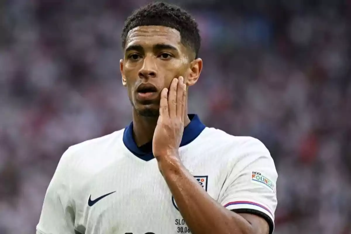 Jugador de fútbol con uniforme blanco tocándose la mejilla en un estadio.