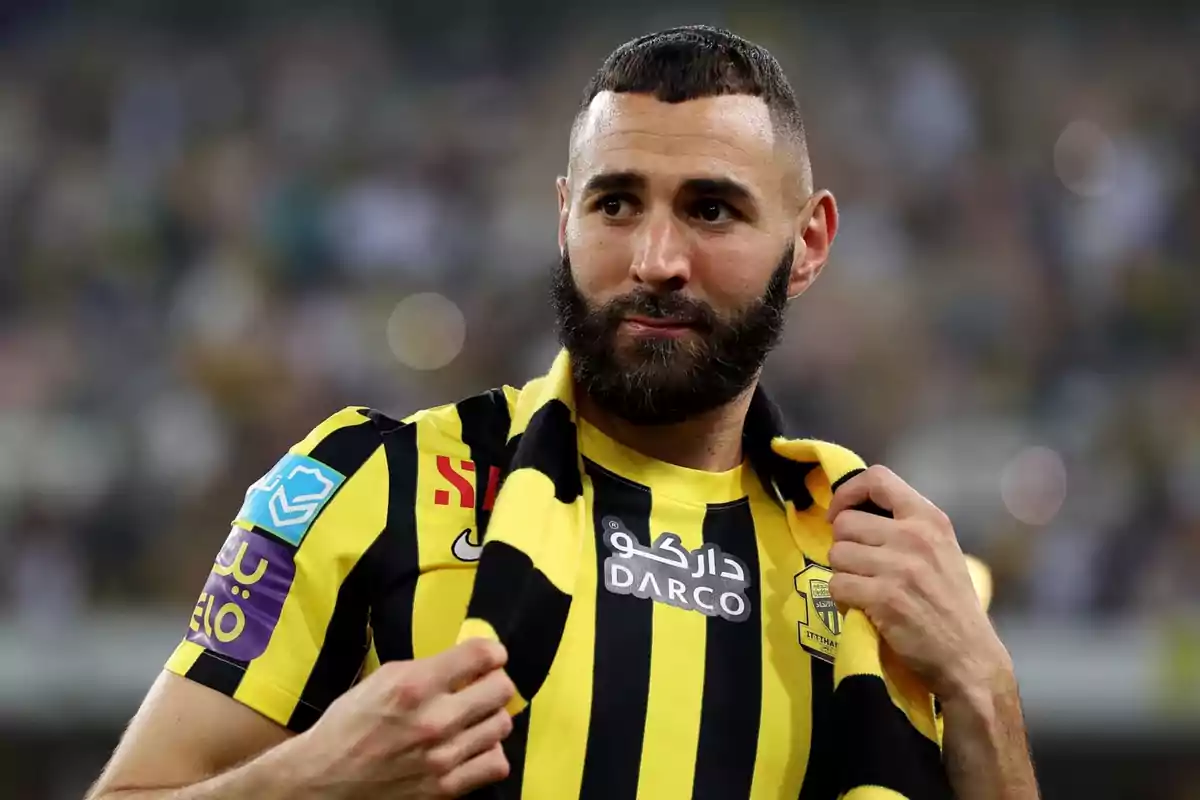 Un jugador de fútbol con barba y cabello corto, vistiendo una camiseta amarilla y negra con logotipos y patrocinadores, sostiene una bufanda del mismo color.