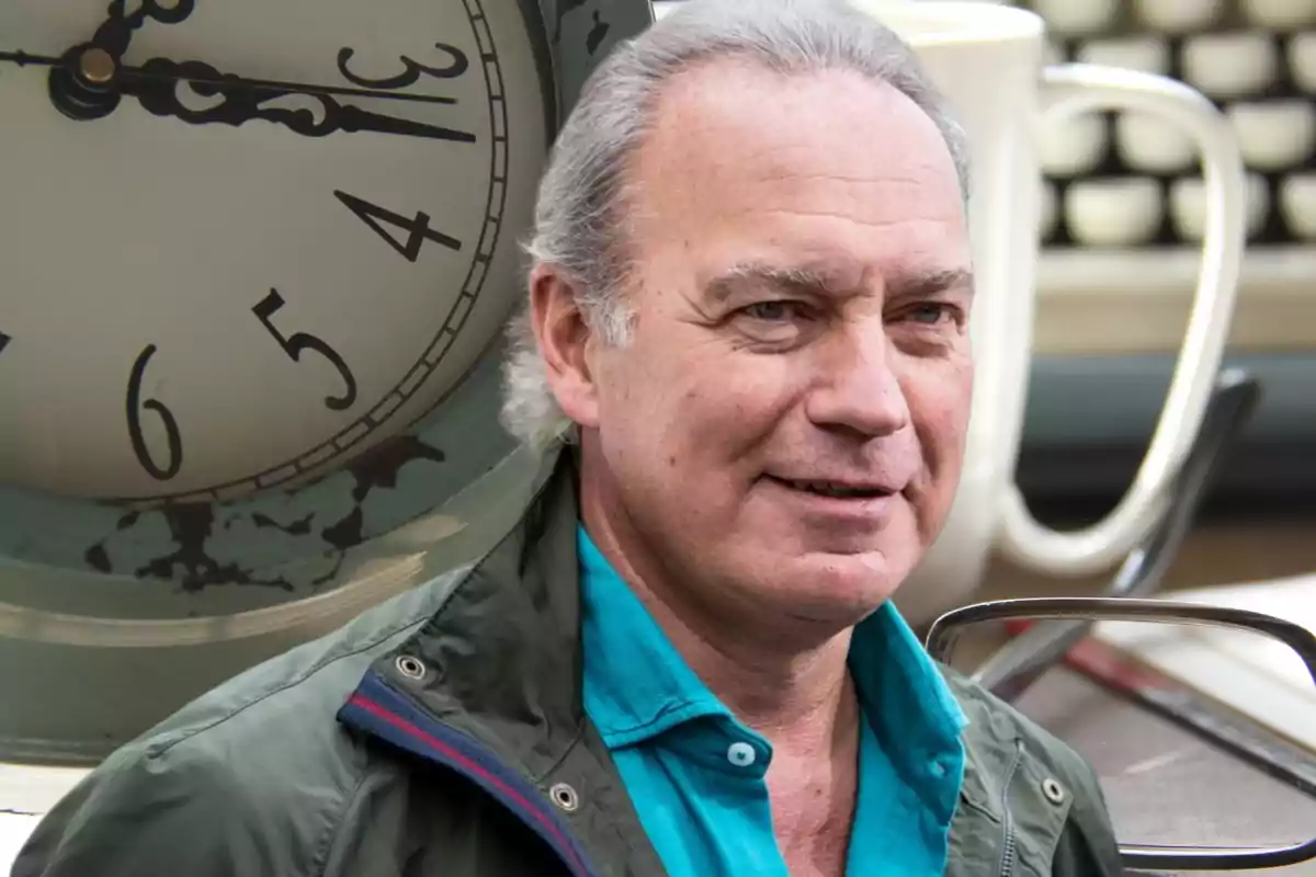 Un hombre de cabello canoso y chaqueta verde está frente a un reloj antiguo y una taza blanca.