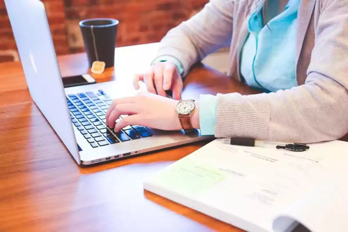 Persona trabajando en una computadora portátil con una taza de café y documentos sobre la mesa.