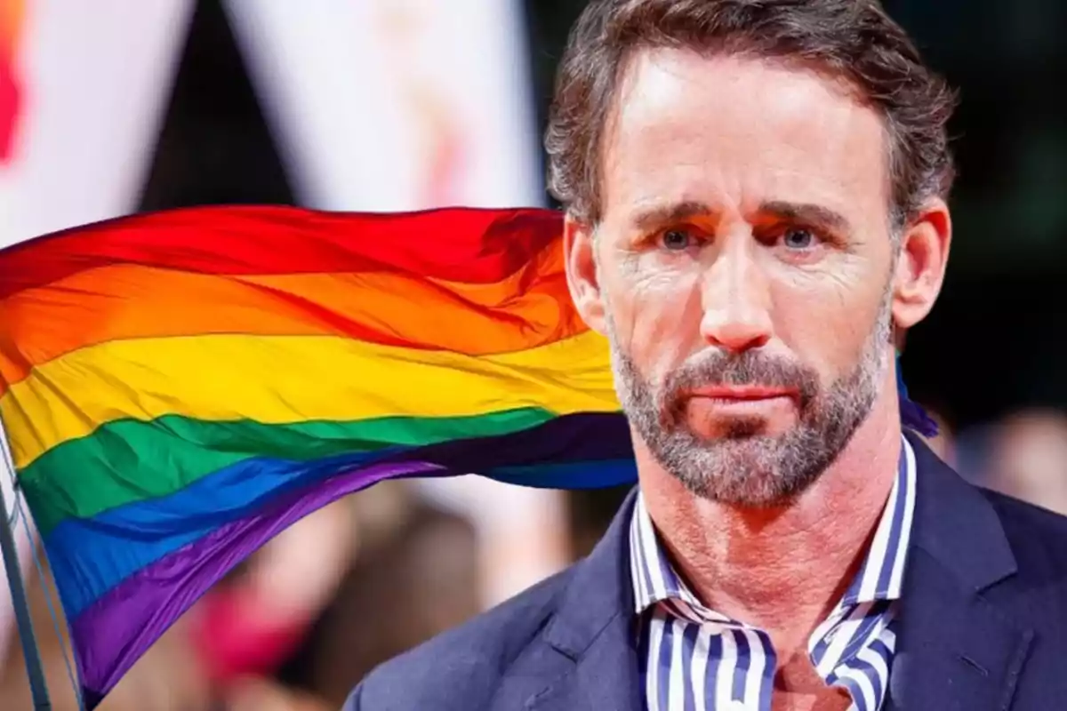 Hombre con barba y cabello corto frente a una bandera del orgullo LGBTQ+ ondeando en el fondo.