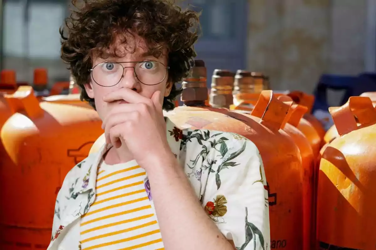 Un joven con gafas y expresión sorprendida se cubre la boca con la mano, mientras está frente a varios cilindros naranjas.