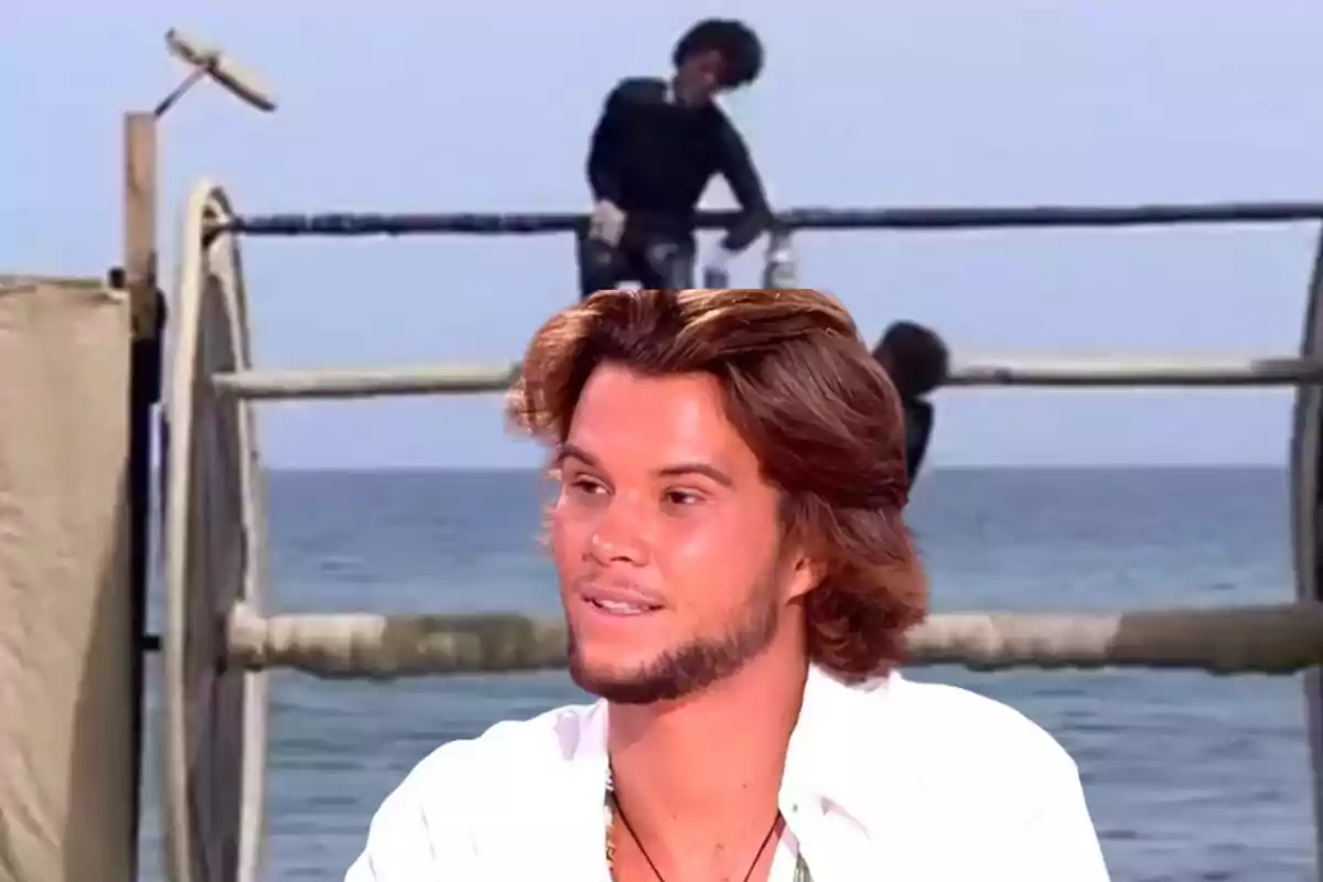 Un hombre con cabello castaño y barba ligera está en primer plano, mientras que en el fondo se ve una estructura de madera y el mar.