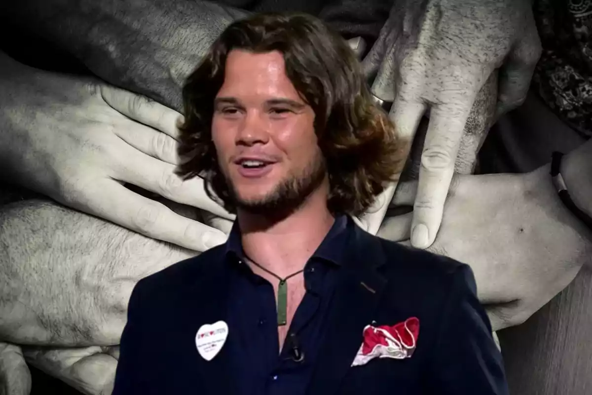 Un hombre con cabello largo y barba ligera, vestido con un traje oscuro y una camisa azul, lleva un pin en forma de corazón en la solapa y un pañuelo rojo en el bolsillo del pecho, con un fondo de manos entrelazadas en blanco y negro.