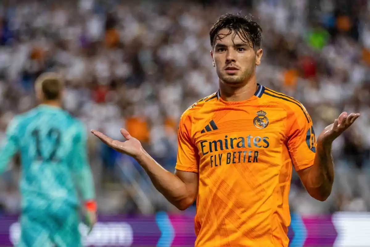 Jugador de fútbol con uniforme naranja del Real Madrid gesticulando con los brazos extendidos en un estadio lleno de espectadores.