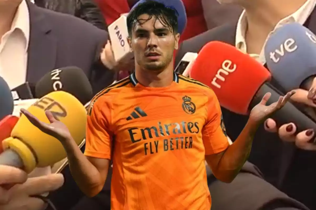 Jugador de fútbol con camiseta naranja del Real Madrid rodeado de micrófonos de prensa.